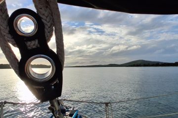 The mainsheet block backlit by sun against blue water and blue sky separated by a thin layer of land