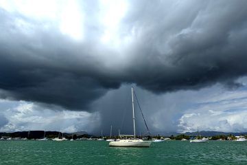 A white-hulled sailboat glows in a sunbeam as a dramatic black cloud dumps a column of rain directly behind it.